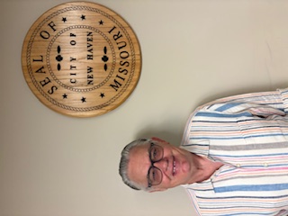 Middle-aged man wearing glasses and a striped shirt standing beside a circular wall seal that reads “SEAL OF THE CITY OF NEW HAVEN, MISSOURI.”